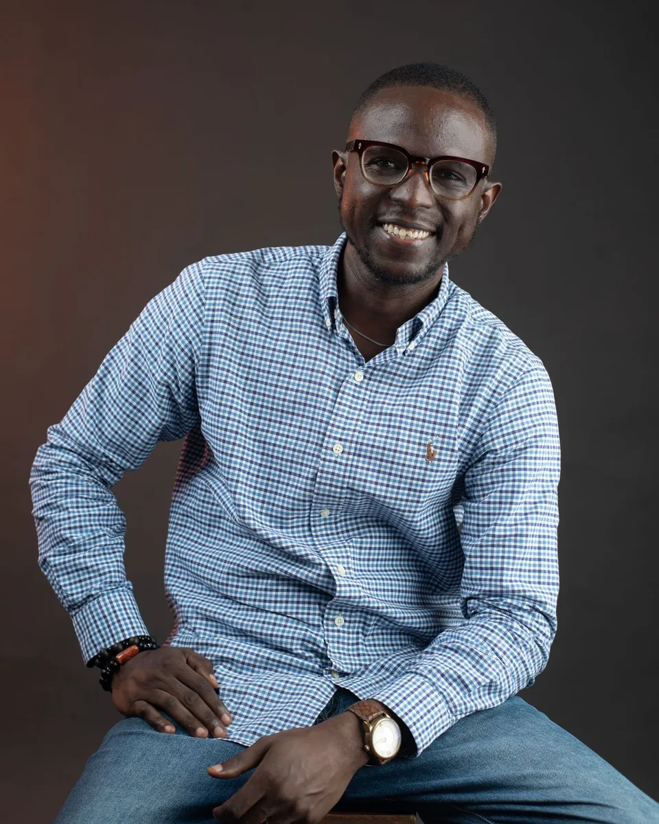 Rotimi Awaye smiling, seated in a blue checked shirt