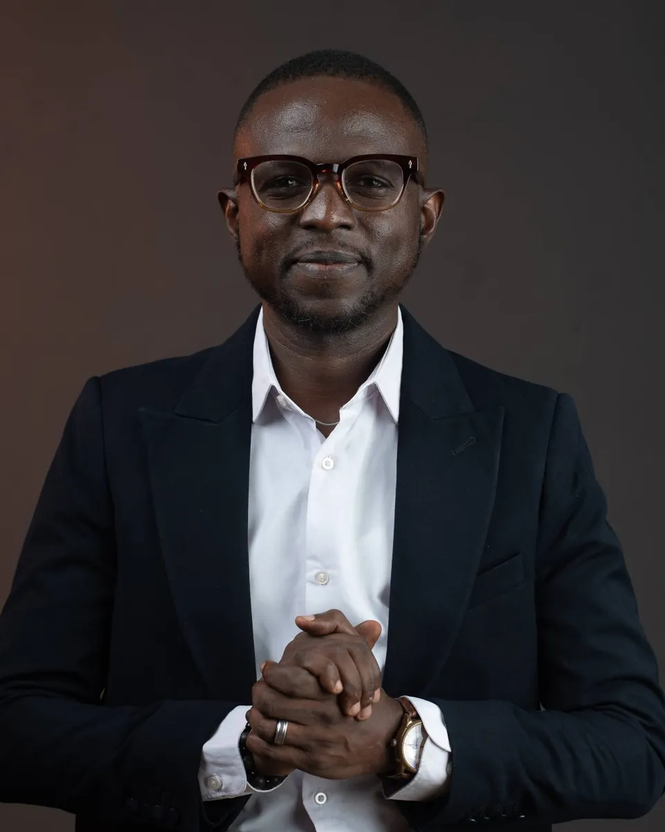 Rotimi Awaye seated in a navy blazer, hands clasped, smiling slightly at the camera